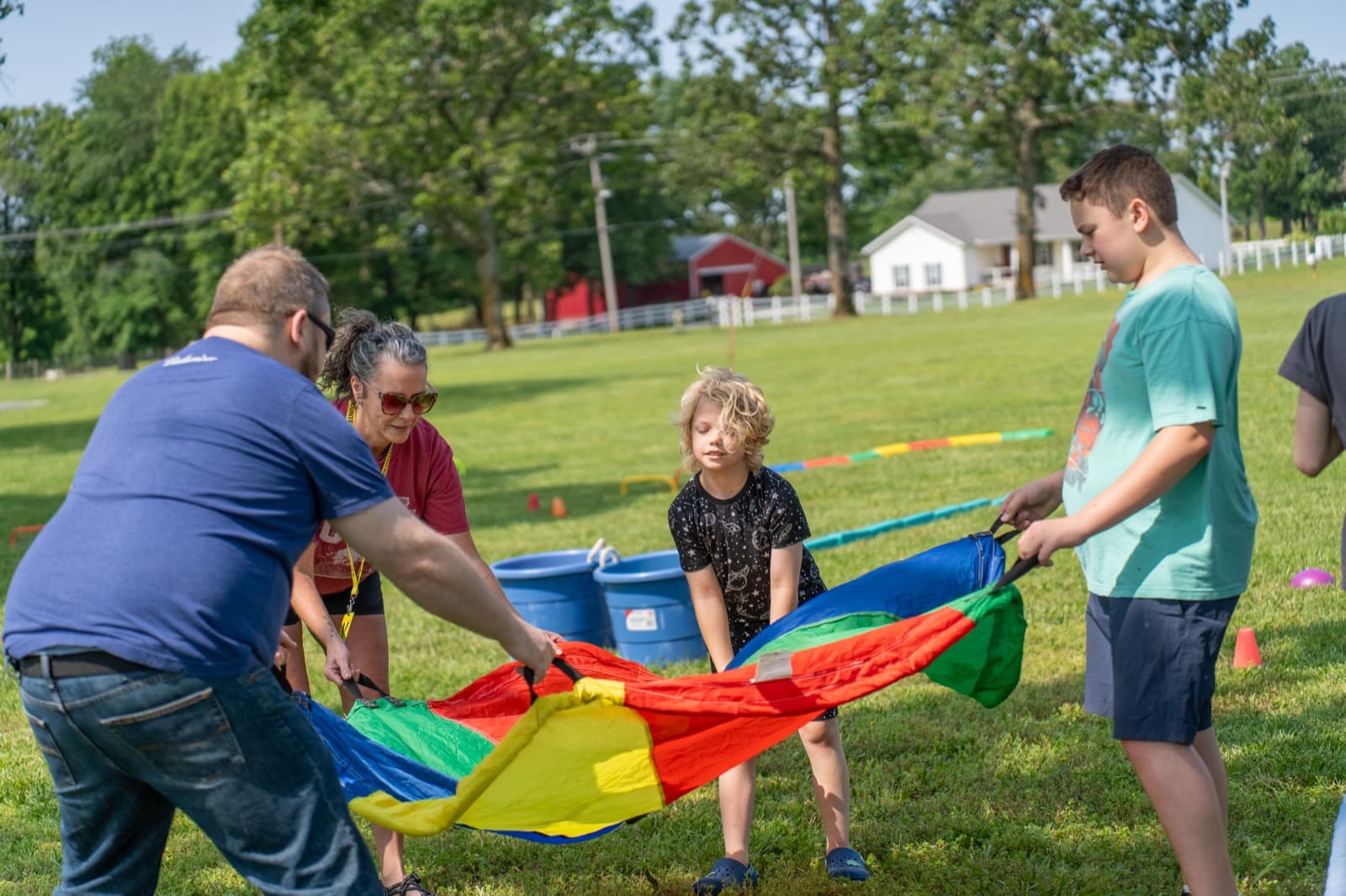 Adults and a child hold the edges of a rainbow parachute together on the grass.