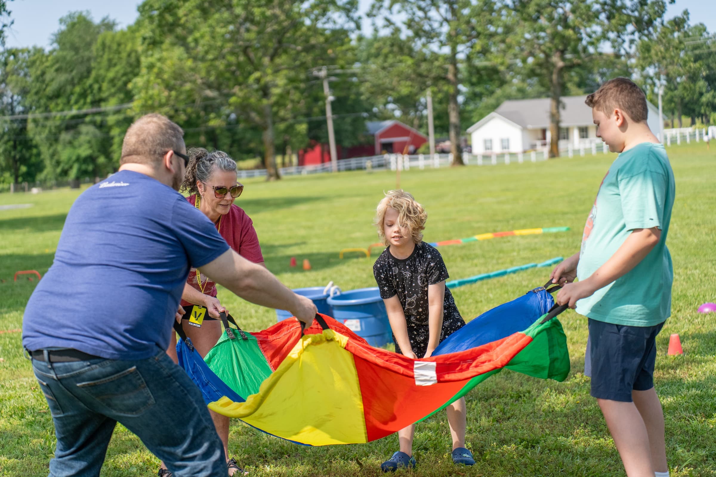 Adults and children together holding out a rainbow parachute on the grass at a community event.