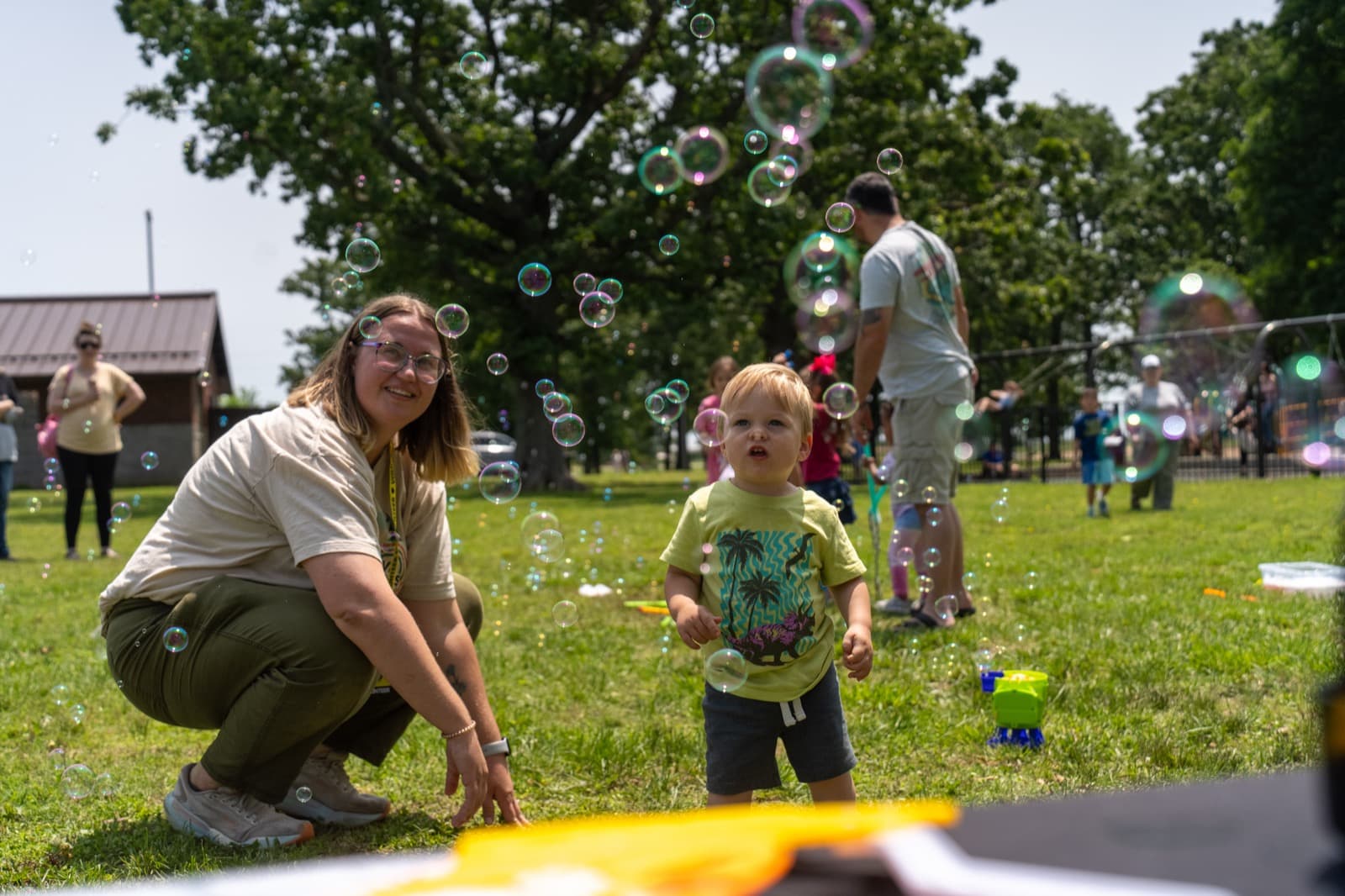 An adult kneels beside a small child as soap bubbles float around them on the grass.